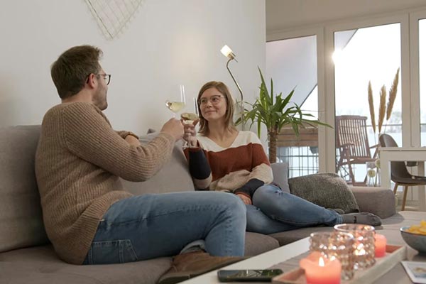 Zwei Personen sitzen auf einem grauen Sofa und stoßen mit einem Glas Wein an